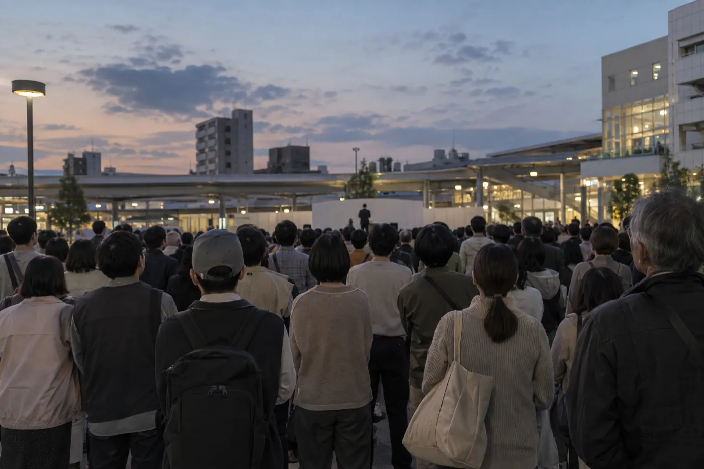 夕方の駅前広場に、大勢の人が後ろ姿で集まっている写真風の画像